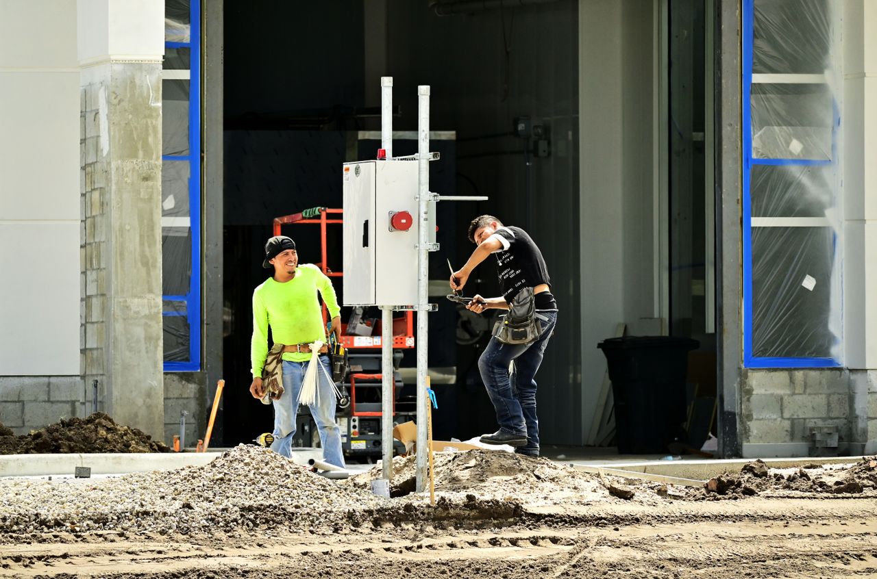 Home Two construction workers install an electrical panel outside a building site, wearing safety gear.